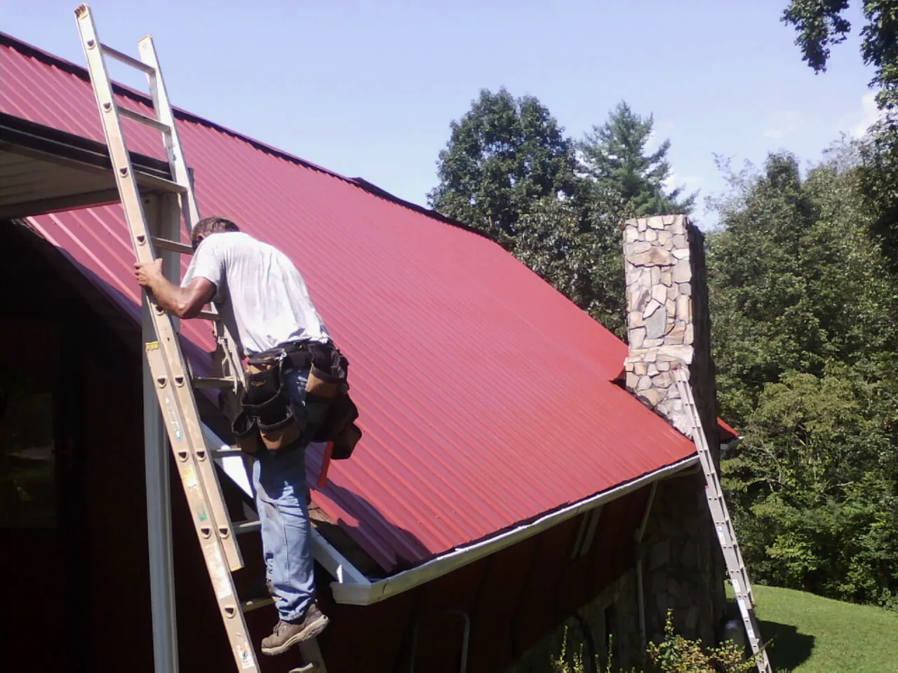 Roofer on a ladder beside a red metal roof and stone chimney — Soffit & Fascia Repair by Roofing Brothers in Lumberville